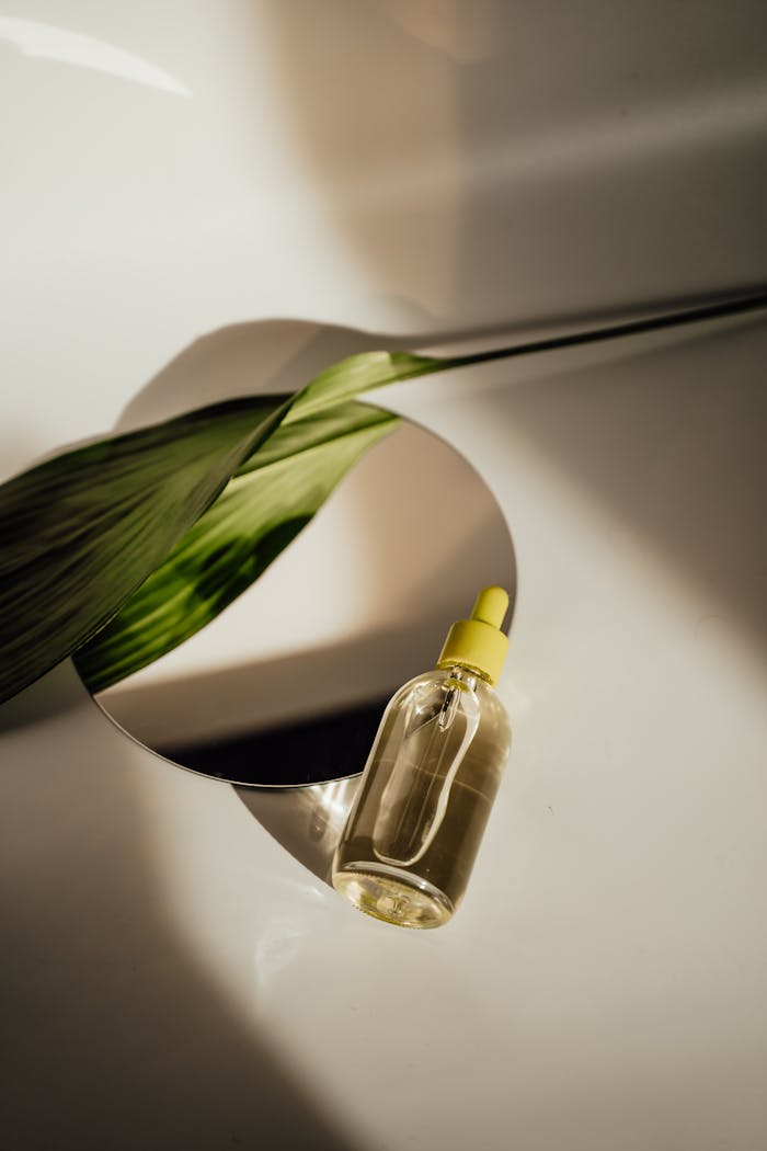 Elegant flat lay of a skincare serum bottle with a leaf reflection on a mirror.